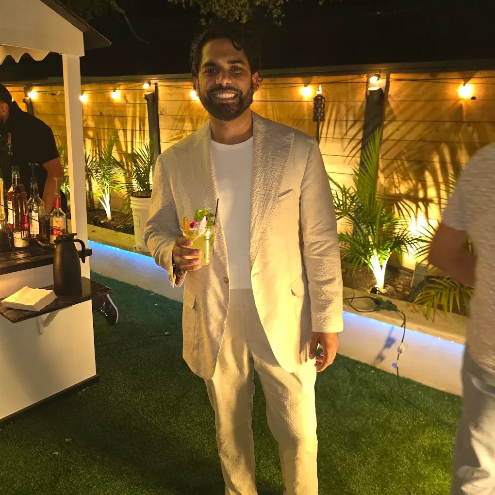 Man in beige suit smiles, holding a drink, standing by a bar outdoors at night.