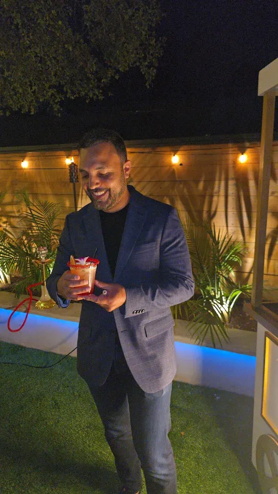 Man in a blue blazer smiles, holding a cocktail outdoors at night with string lights and greenery.
