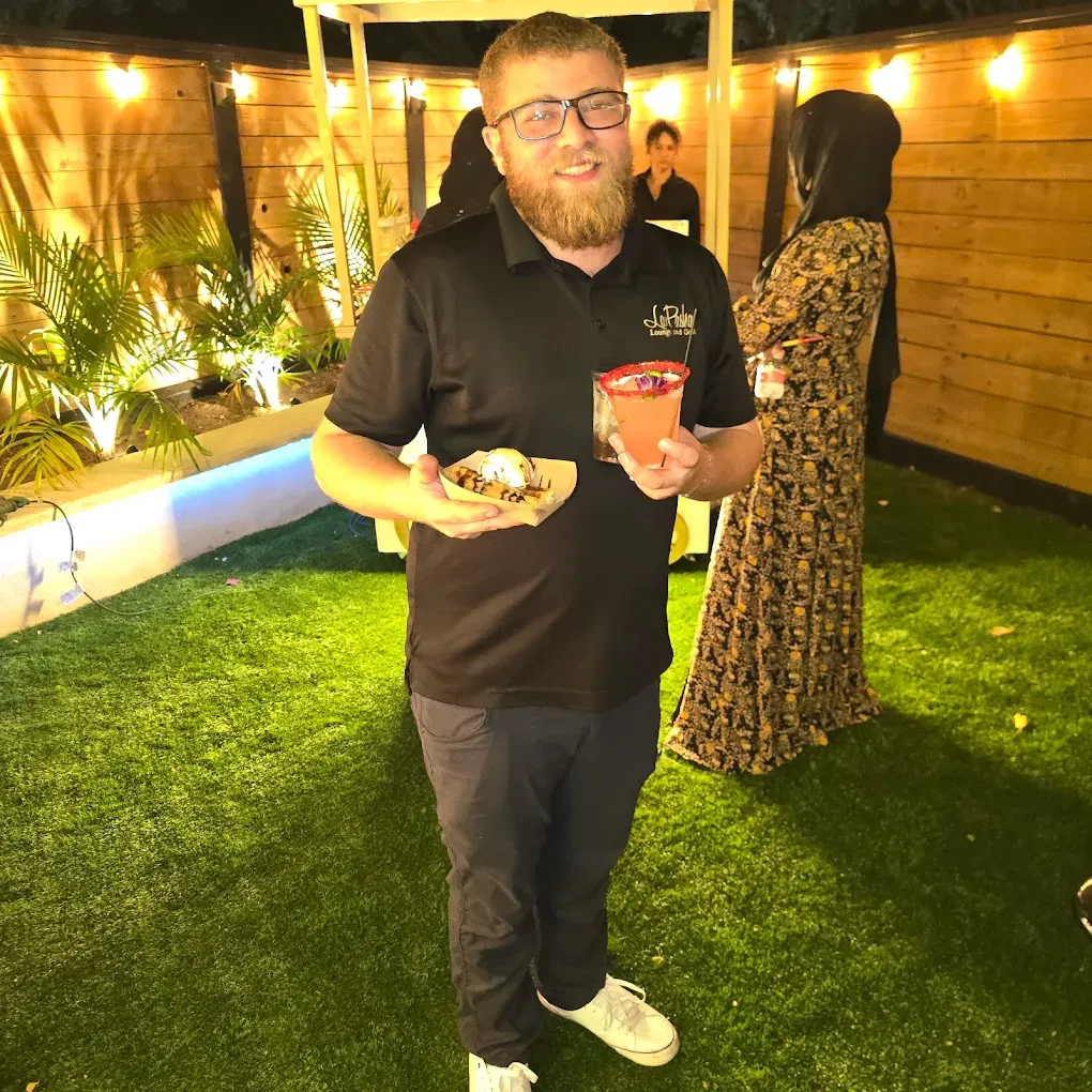 Man holding food and drink, smiles at camera in an outdoor setting with guests.