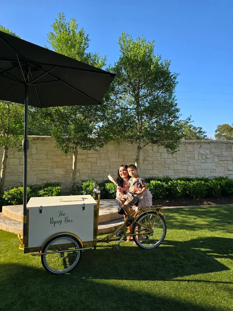 People beside ice cream cart with umbrella, green lawn, trees, and stone wall in background.