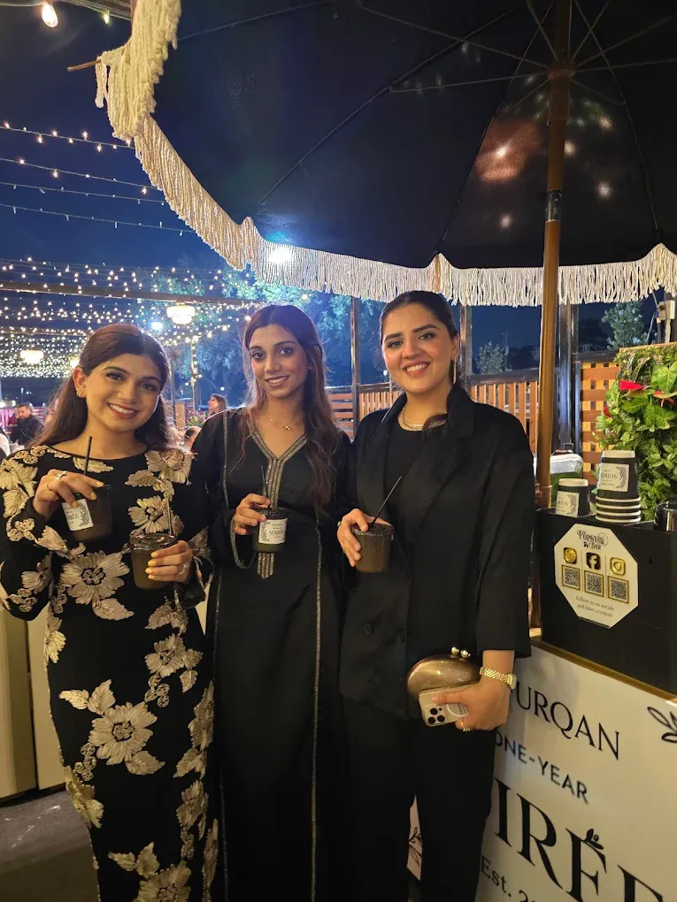 Three women holding drinks, posing outdoors at night, under lights.