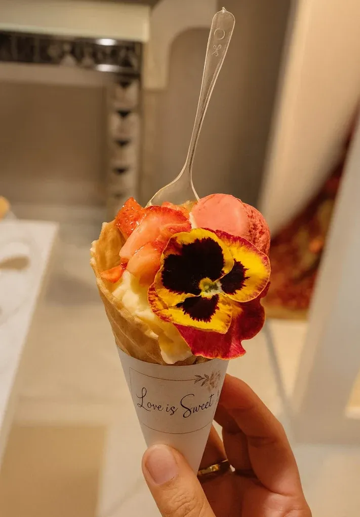 Ice cream cone with scoops, strawberries, and a flower, held in a hand.