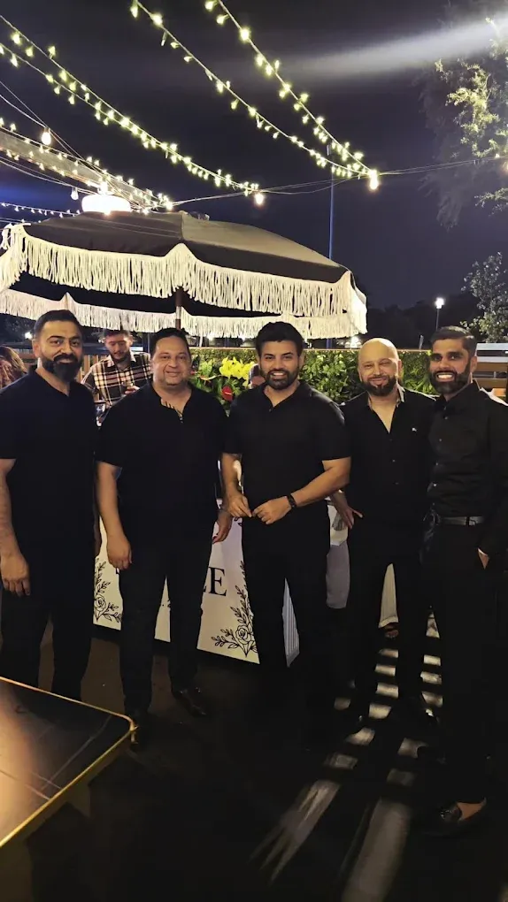 Five men in black outfits pose outdoors at night, under string lights, near a decorative structure.