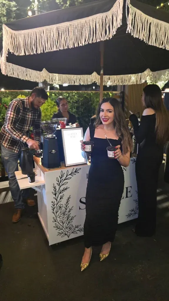 Woman in black dress and gold heels at an outdoor bar with drinks, others visible.