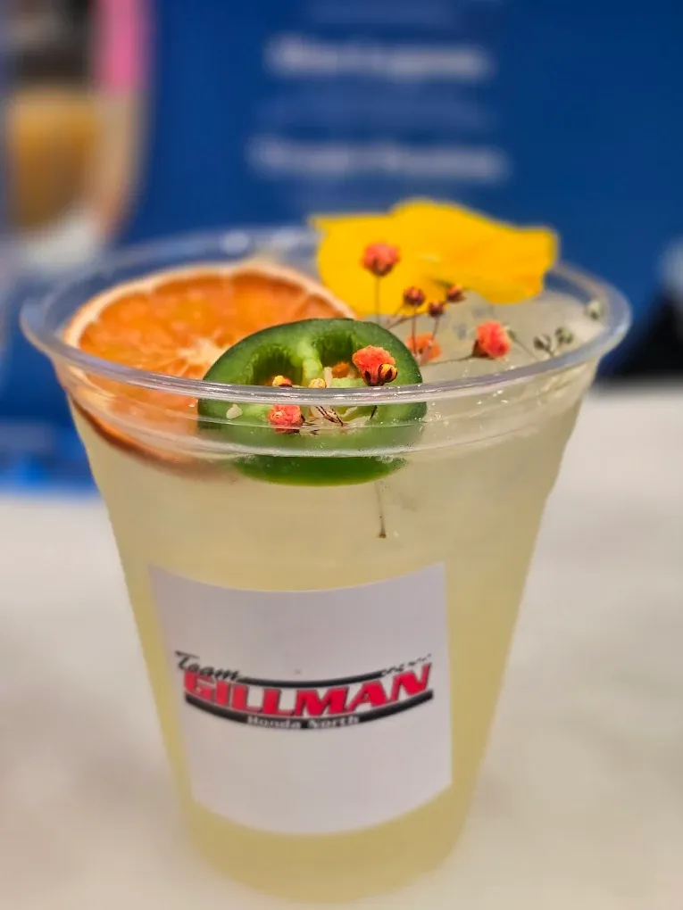 A clear plastic cup of margarita with jalapeño, orange slice, mango, and flower garnish. Team Gillman logo.