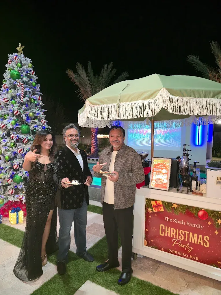 Three people pose at an outdoor Christmas party; white tree with decorations and bar cart in background.