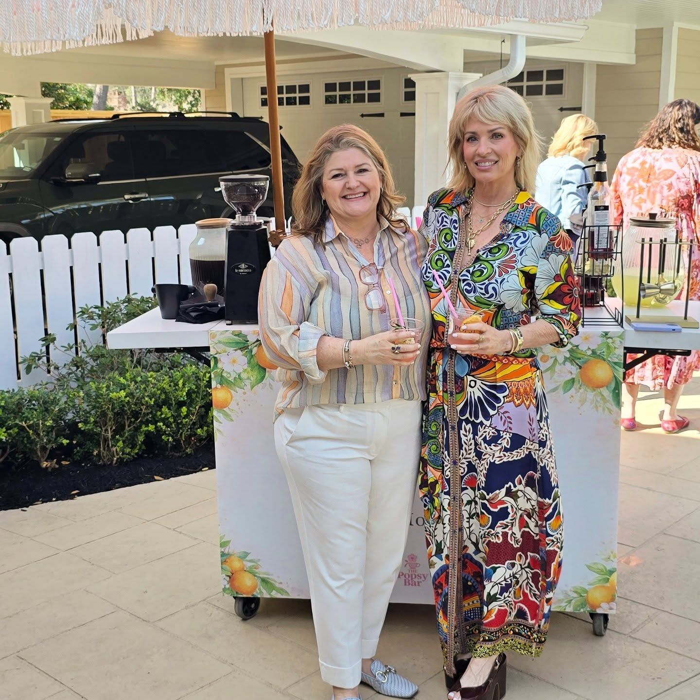 Two people pose in front of a portable beverage bar at an outdoor event near a white fence and vehicle.