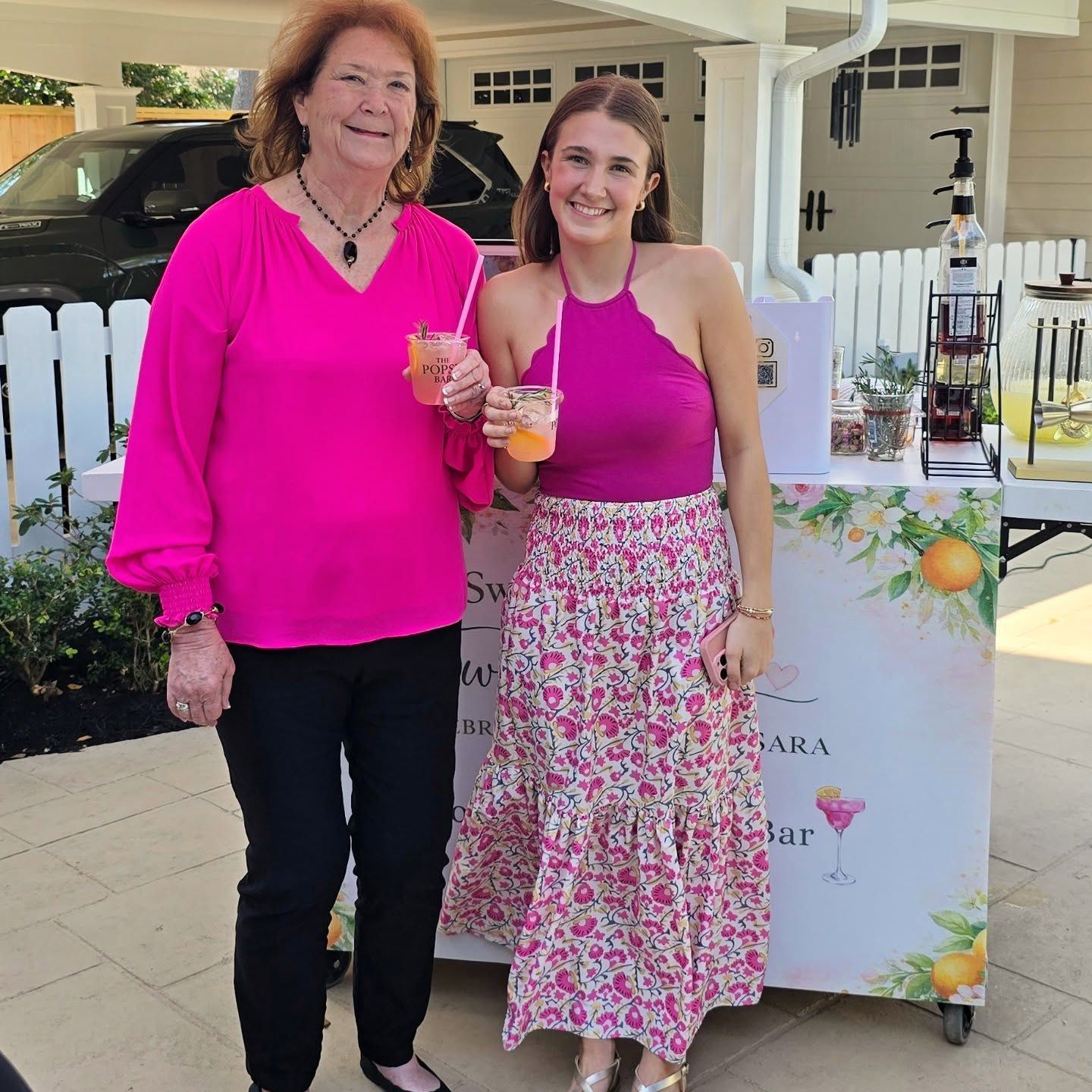 Two women smiling, holding drinks, standing by a beverage cart, matching pink outfits.