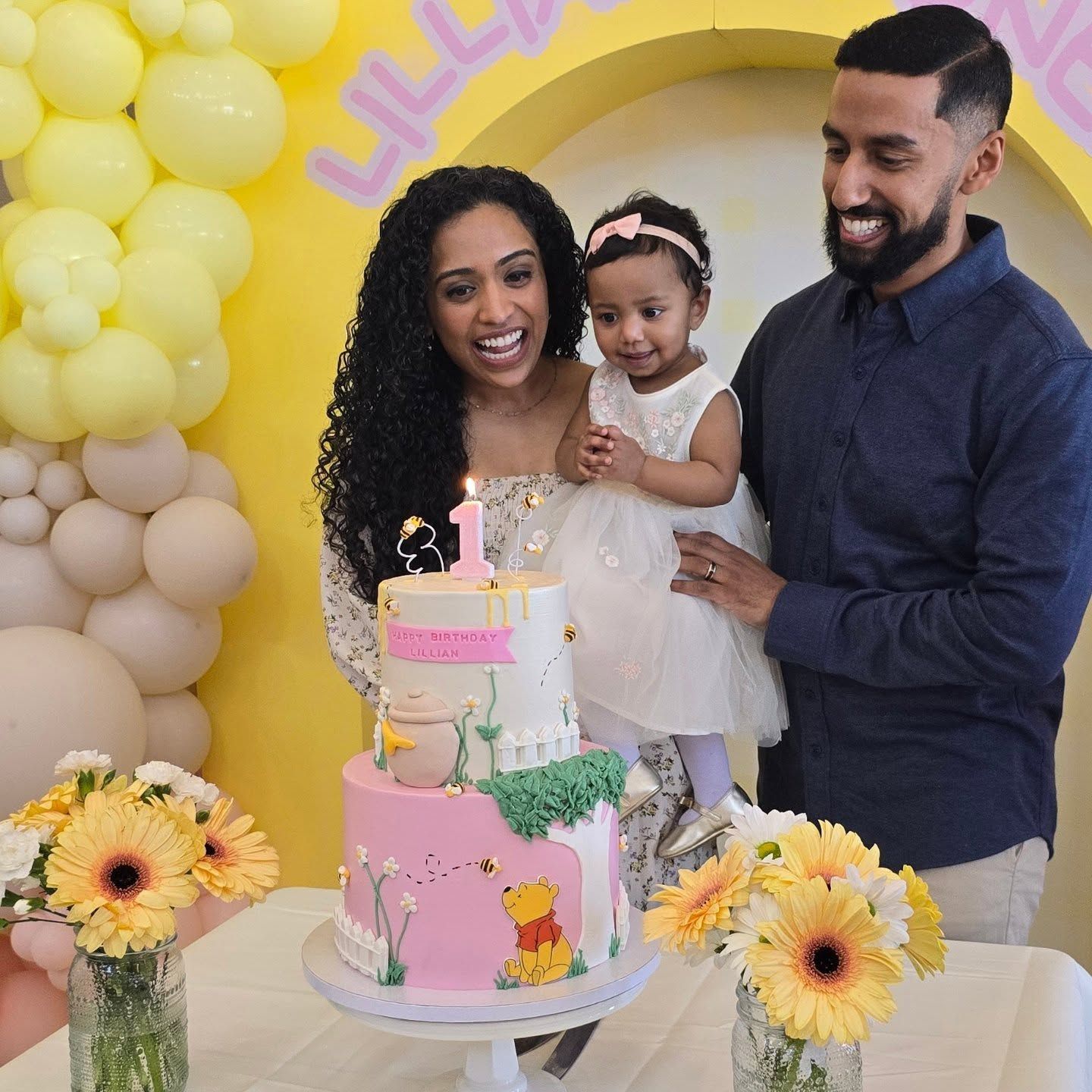 Family celebrating a child's first birthday with cake, flowers, and yellow balloon decorations. They appear joyful.