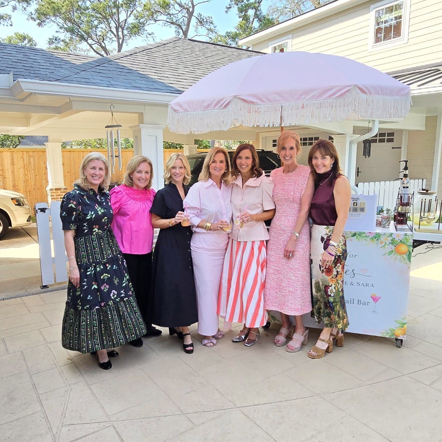 Seven women pose around a light blue bar with a pink umbrella in an outdoor setting.