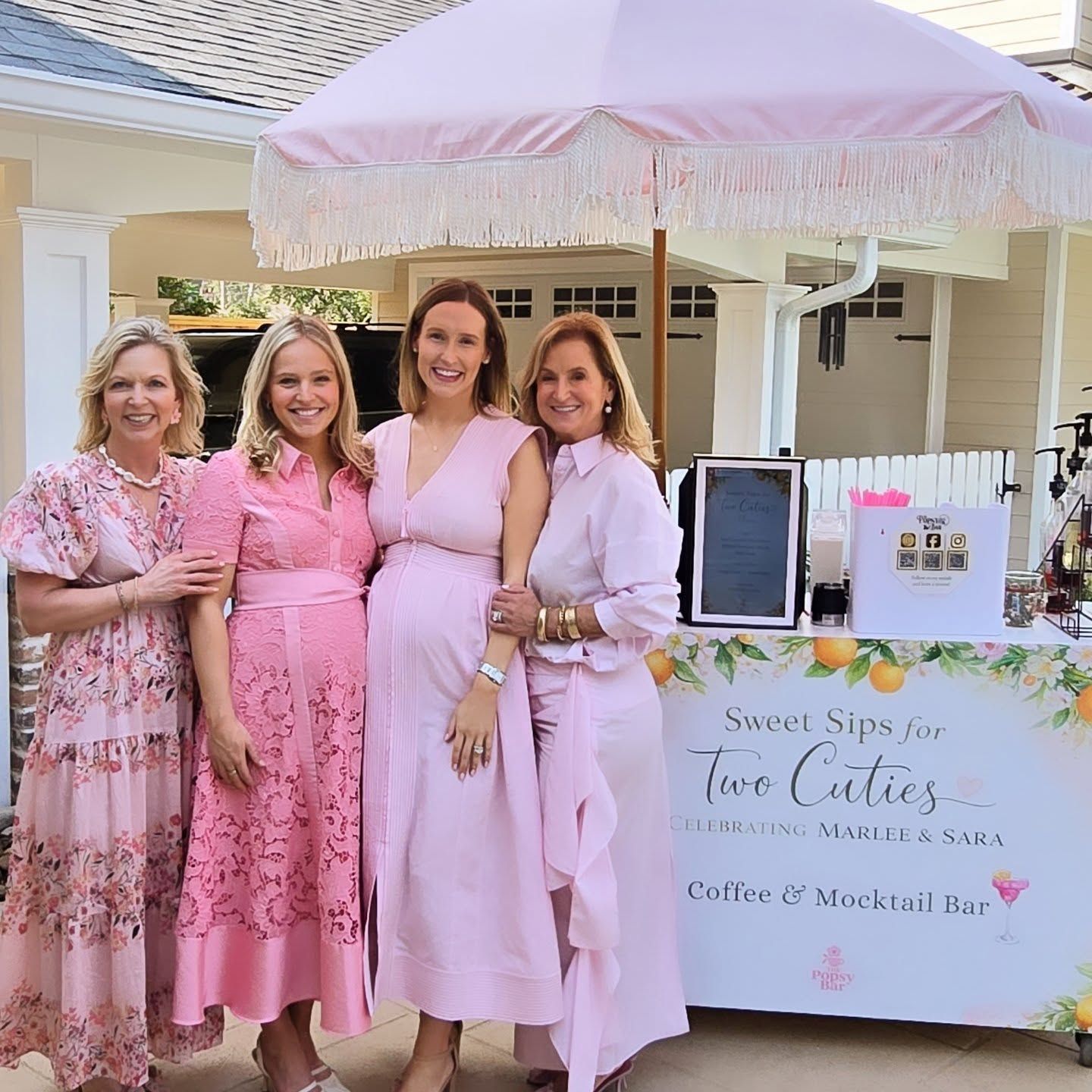 Four women in pink outfits pose by a pink-themed drink stand with an umbrella in a sunny outdoor setting.
