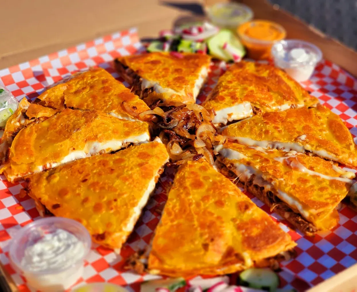 Large, golden-brown quesadilla cut into wedges, on a checkered red-and-white surface, with dipping sauces and salad.