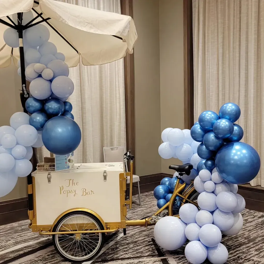 A white cart with an umbrella, adorned with clusters of light blue and metallic blue balloons, parked on a carpeted floor.