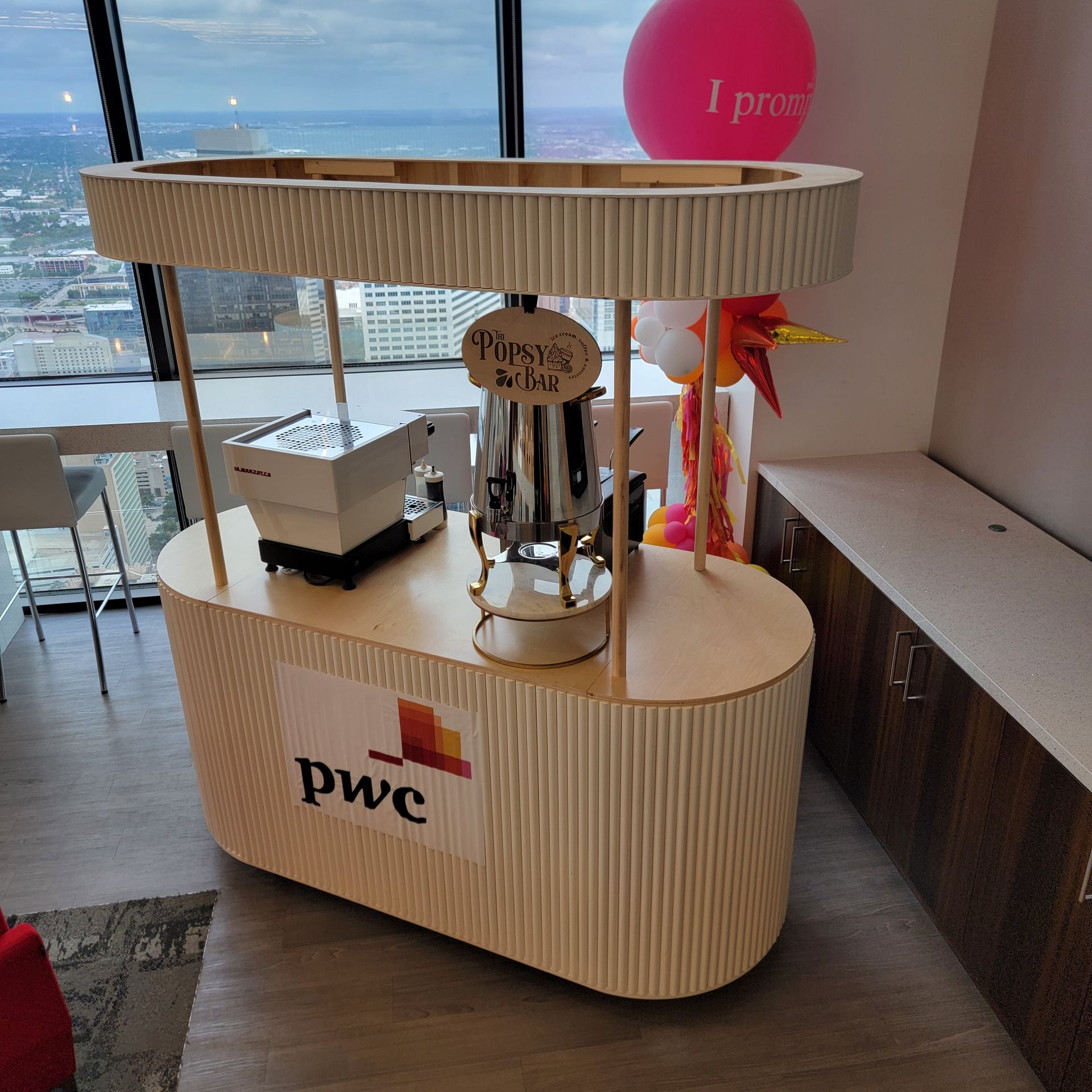 A mobile light-wood coffee station with a PwC logo, an espresso machine, and a metal server, located in a high-rise office.