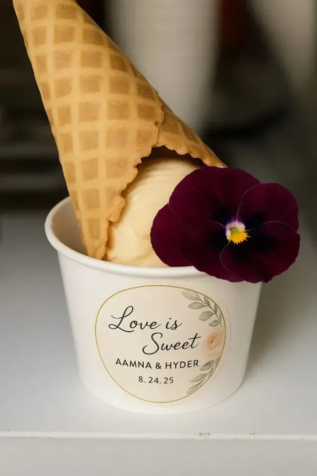 A small cup of ice cream with a waffle cone, garnished with a deep purple flower, labeled for a wedding on 8.24.25.