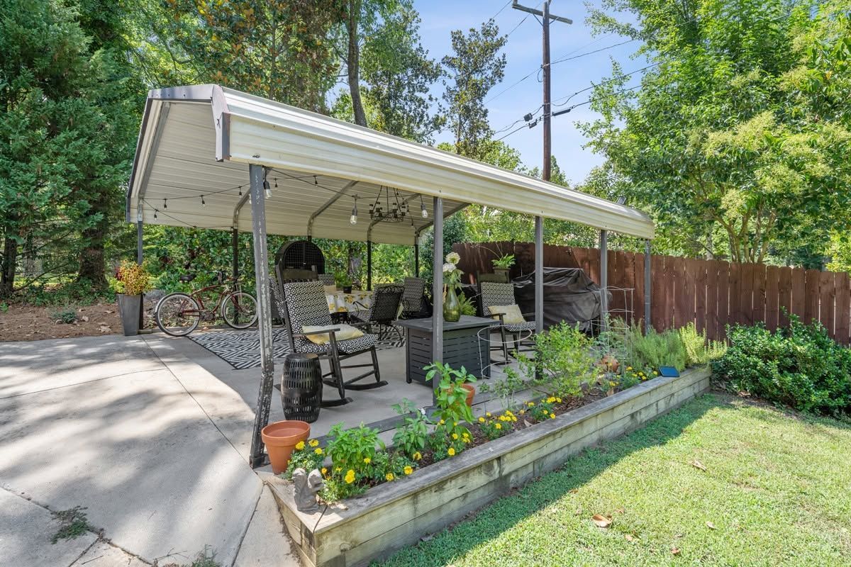 Covered patio with outdoor seating beside a garden bed and wooden fence in a sunny backyard.