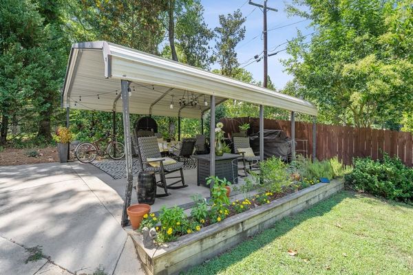 Covered backyard patio with chairs, potted plants, raised flower bed, and a wooden fence beside a lawn
