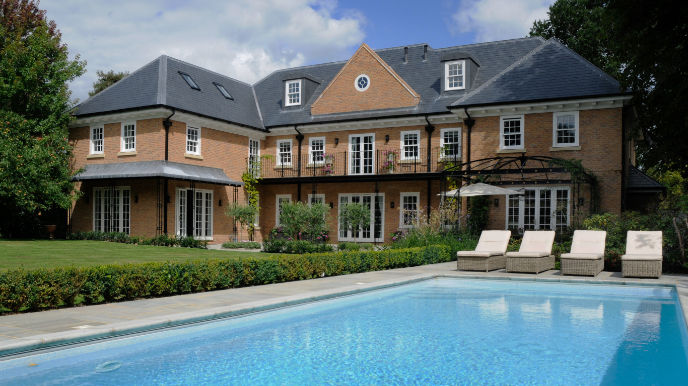 Large brick house with a pool, patio chairs, and hedges under a partly cloudy sky
