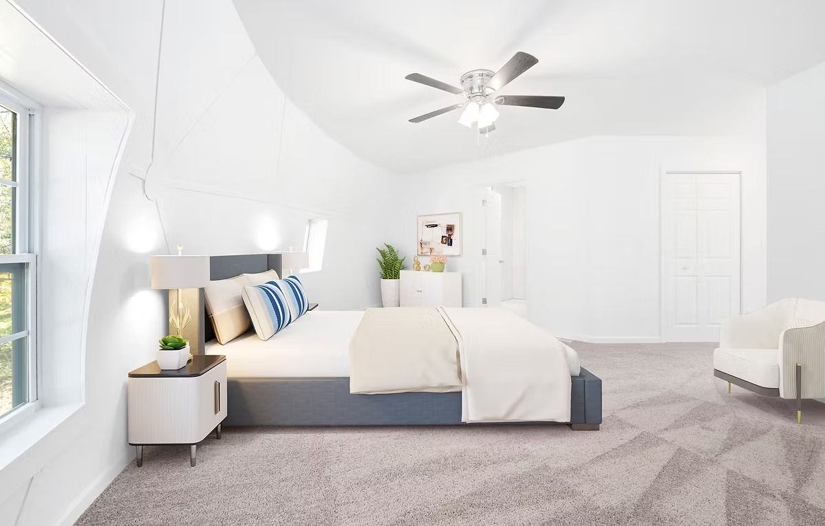 Bright white bedroom with gray carpet, neatly made bed, windows, ceiling fan, and an armchair.