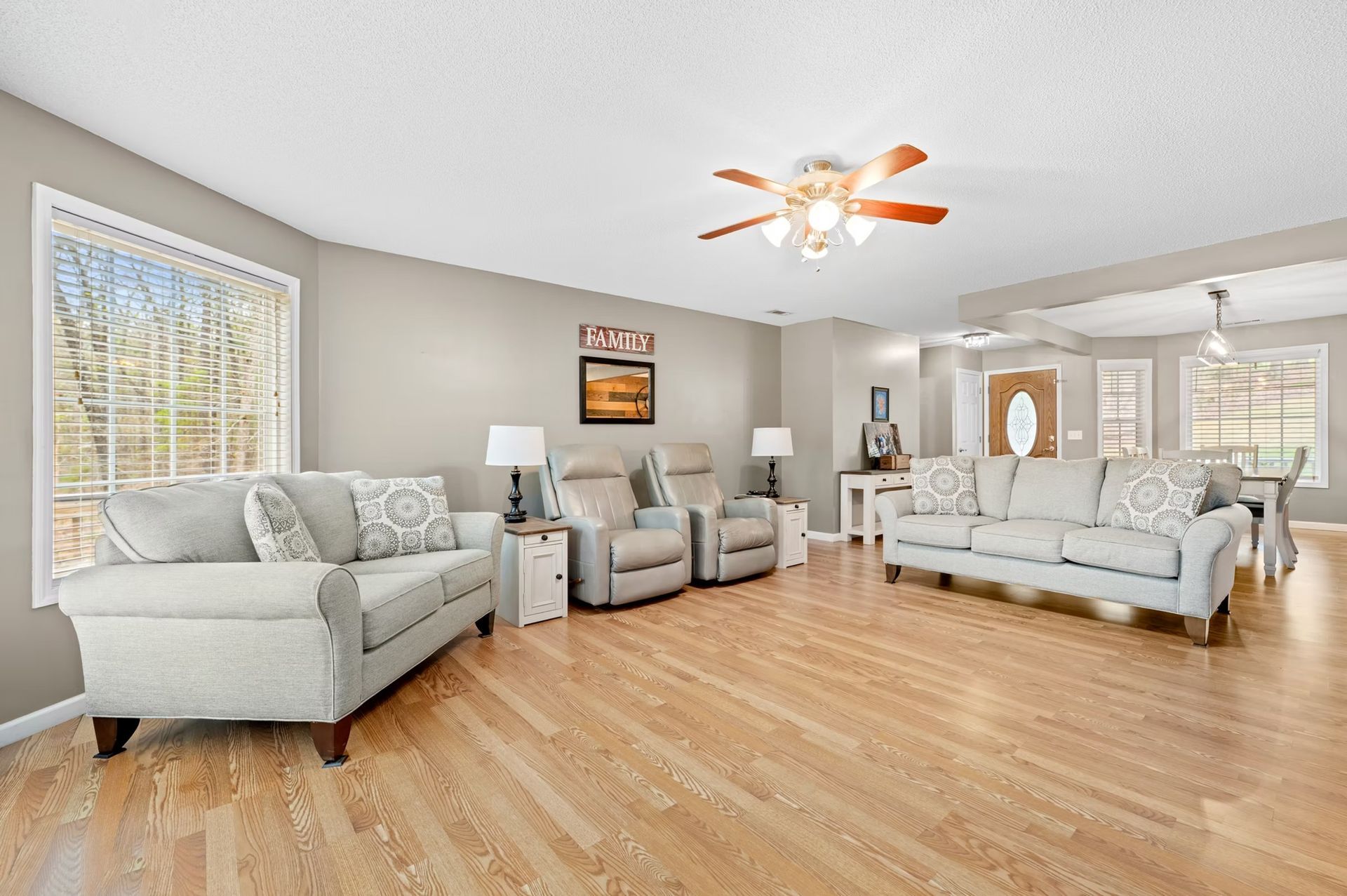 Bright living room with gray sofas, recliners, wood floors, and a ceiling fan near large windows.