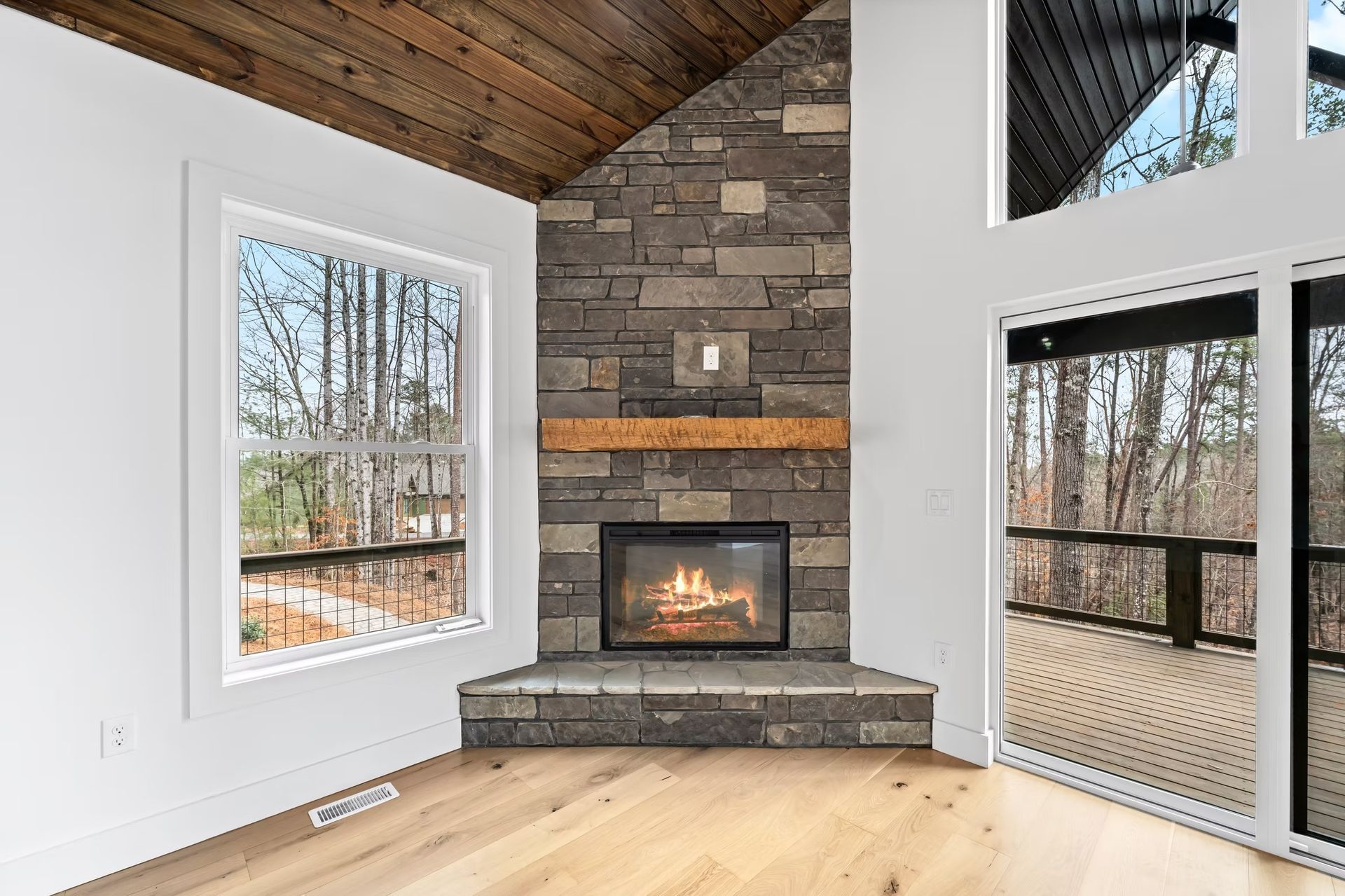 Modern living room with stone fireplace, wood ceiling, and large windows overlooking trees