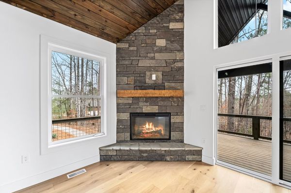 Modern living room with stone fireplace, wood ceiling, and large windows overlooking trees
