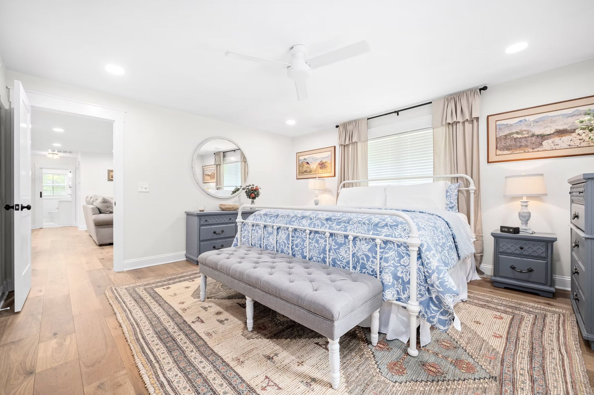 Bright bedroom with blue bedspread, bench, dresser, and patterned rug in a light, airy room