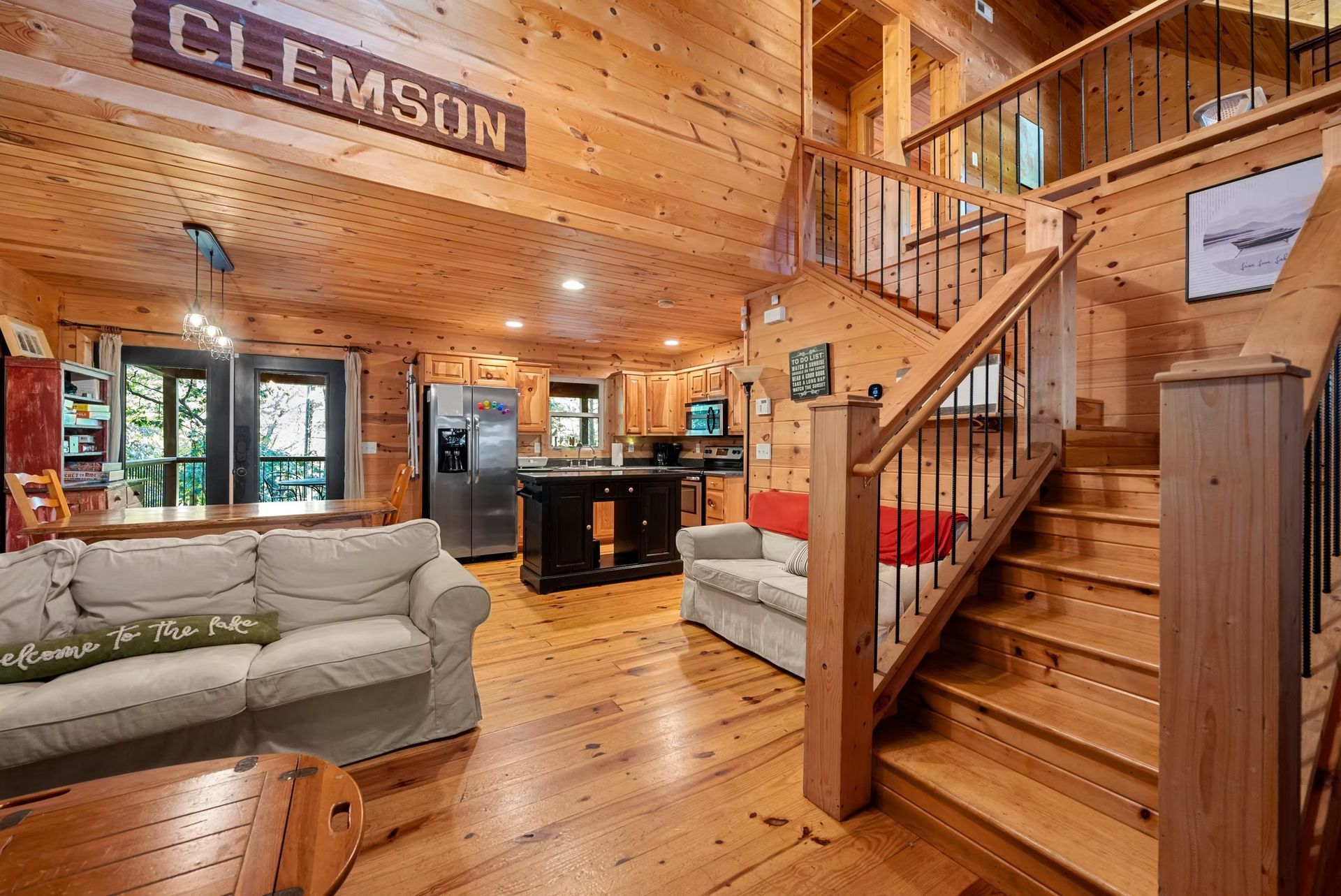 Cozy log cabin living room with wood walls, staircase, gray sofa, and “CLEMSON” sign above the doorway