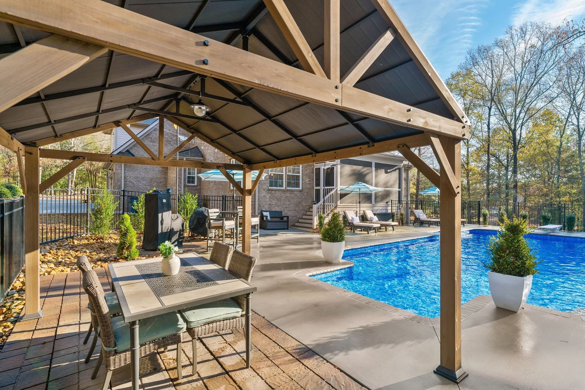 Covered patio beside a blue swimming pool with dining table and potted plants on a sunny day