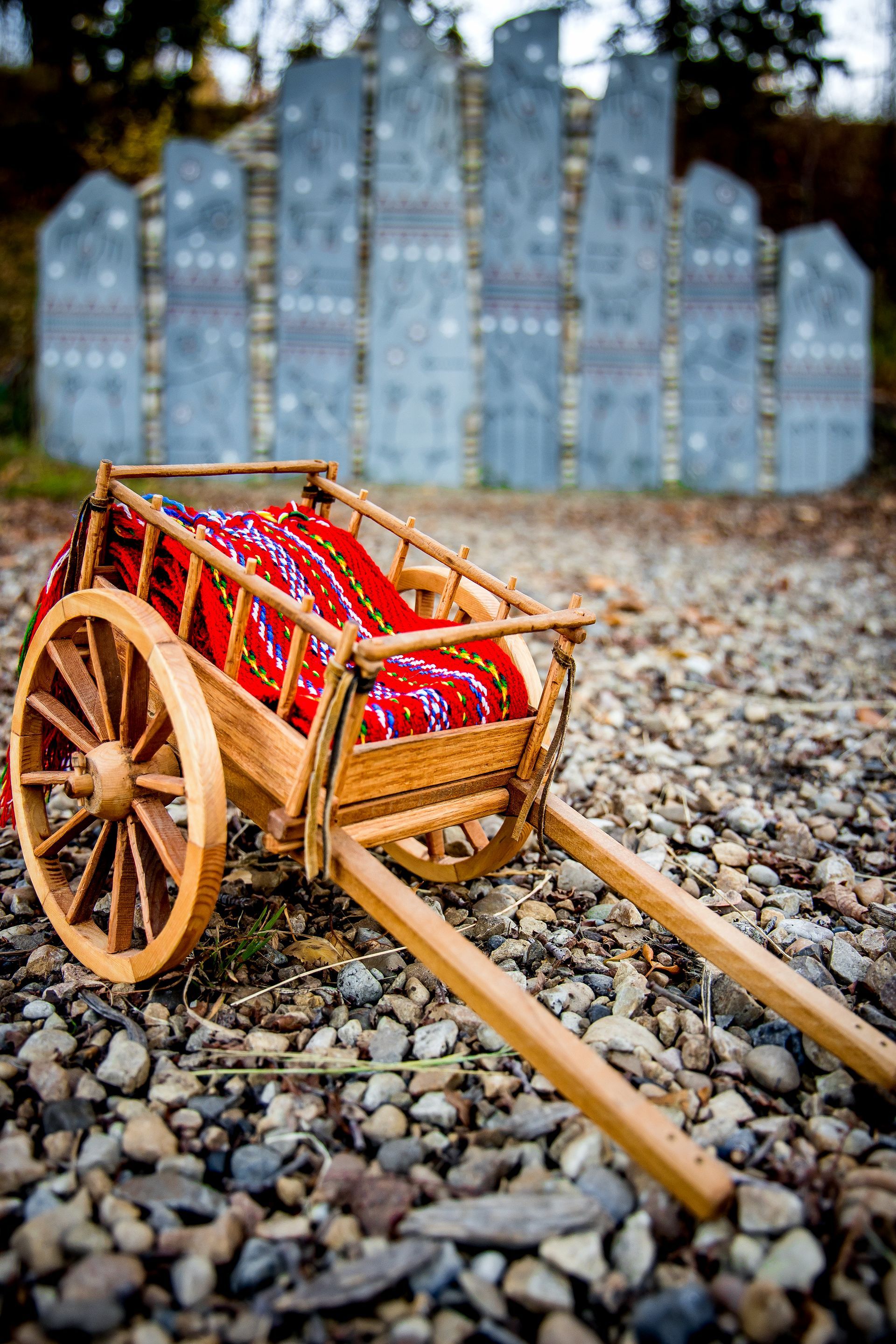 Wooden Red River Cart Replica with a Metis sash in front of a Cree monument in an Indigenous park