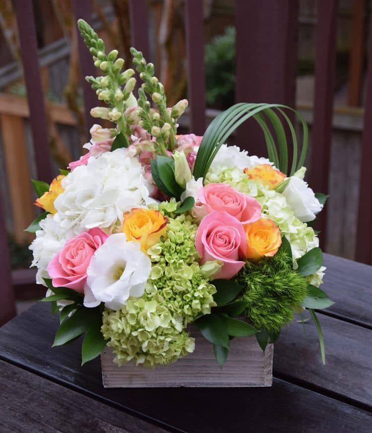 Floral arrangement in a wooden box featuring pink, yellow, and white roses, green hydrangeas, and snapdragons.
