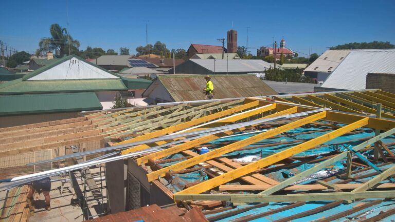 A construction worker on a roof under repair, with exposed wooden trusses, blue underlay, and a town backdrop. — Daniher Building Company in Flynn, ACT