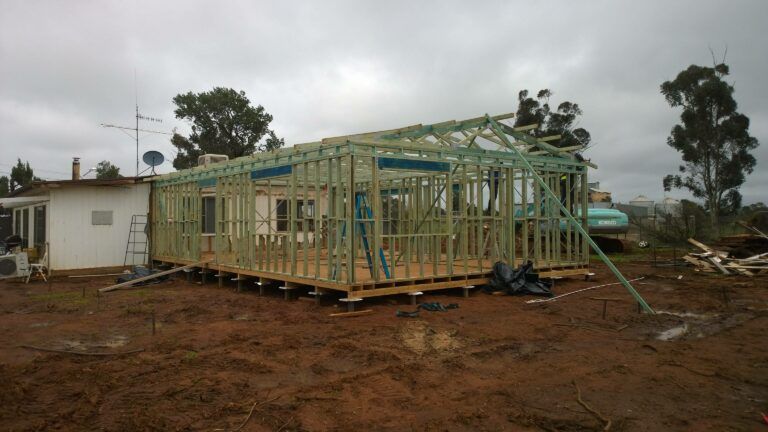 A wooden wall and roof frame for a house addition being built next to an existing white home under a cloudy sky. — Daniher Building Company in Flynn, ACT
