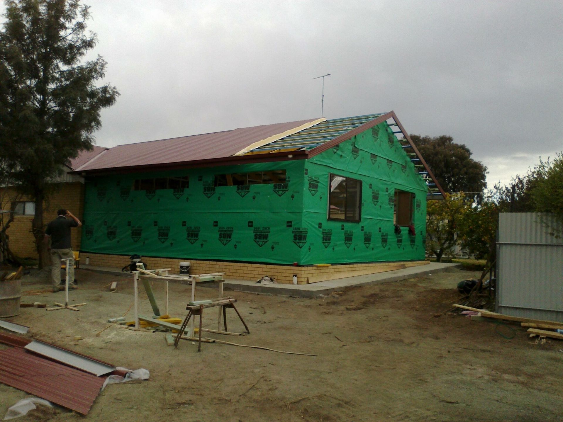 A person stands by a house exterior under construction, wrapped in green protective sheeting with a partially tiled roof. — Daniher Building Company in Flynn, ACT