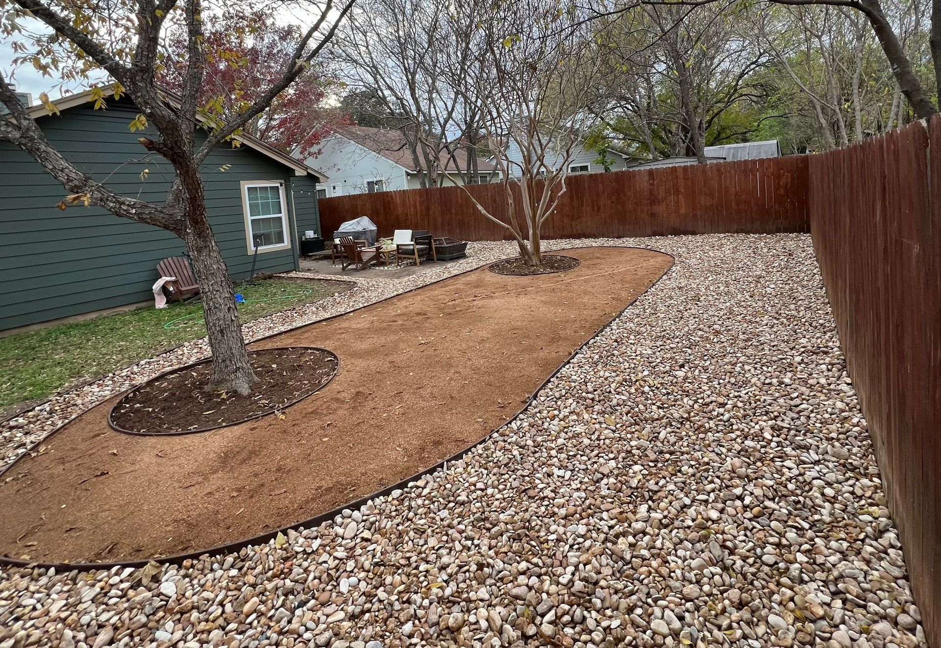 Backyard landscaping with gravel and mulch paths, surrounded by a wooden fence and trees.