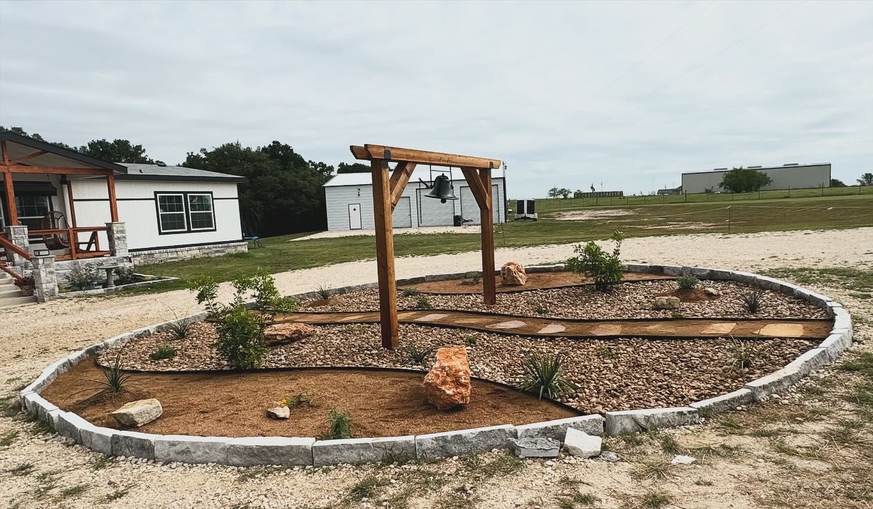 Oval garden bed with wooden arch, stone border, mulch, and small plants; gravel path.