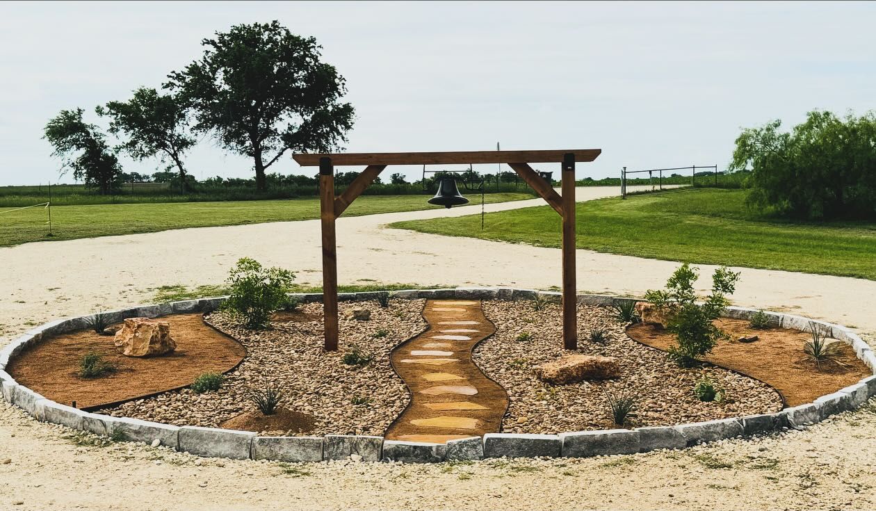 Wooden archway in a circular garden with a stone path, surrounded by gravel and mulch, set in a grassy field.