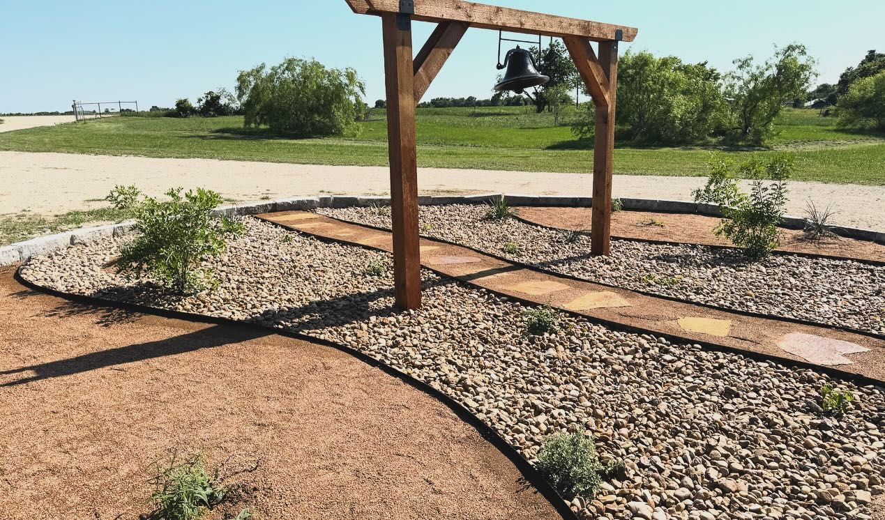 Wooden archway over a gravel path with stepping stones, surrounded by plants and brown mulch, in a sunny outdoor setting.
