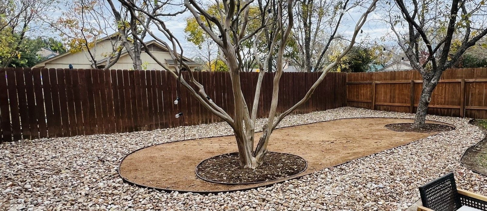 Backyard with a brown wooden fence, trees, gravel pathway, and a central dirt area with trees.
