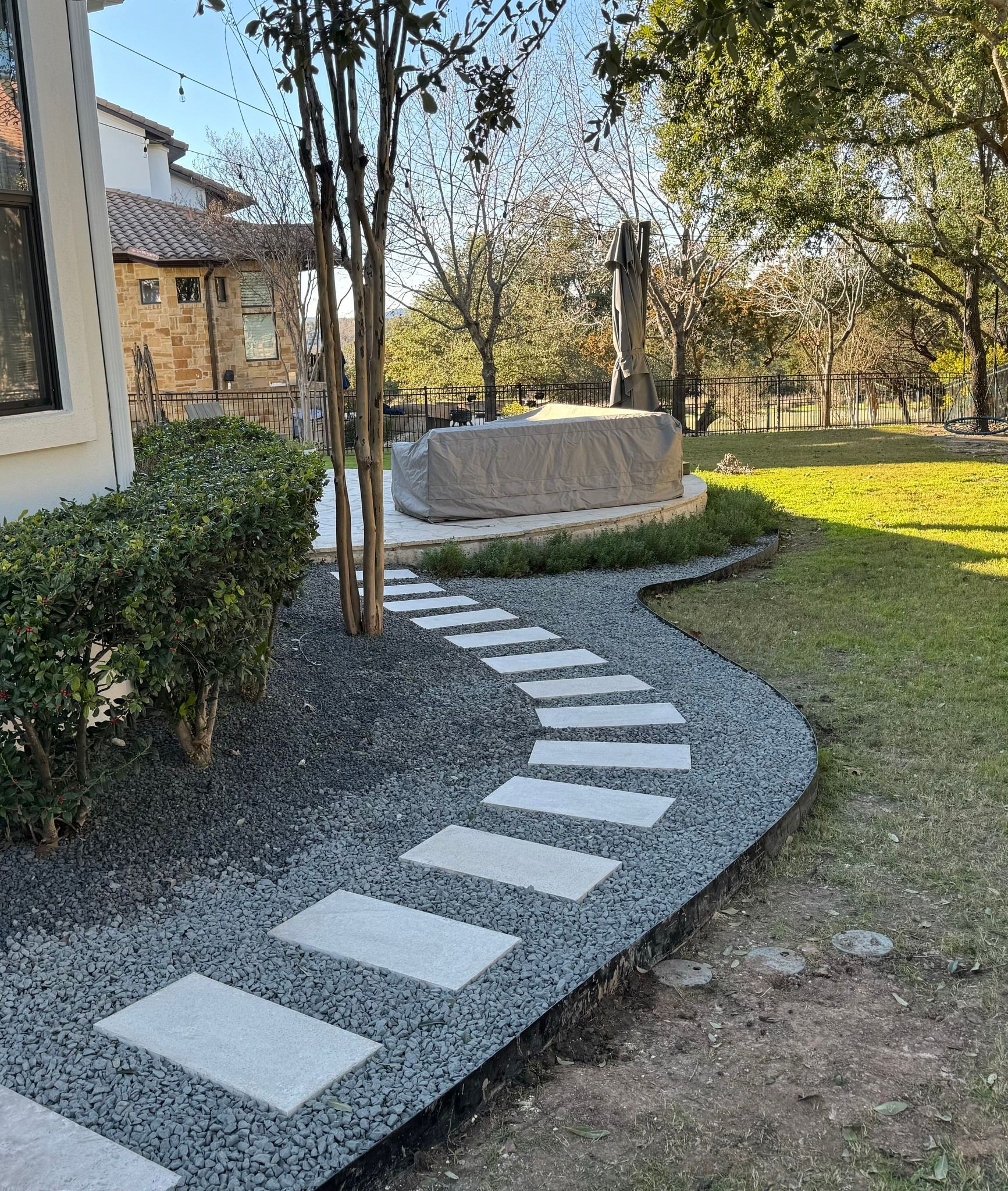 Stone path curves through gravel, leading to a large stone block in a grassy yard, near a building and trees.