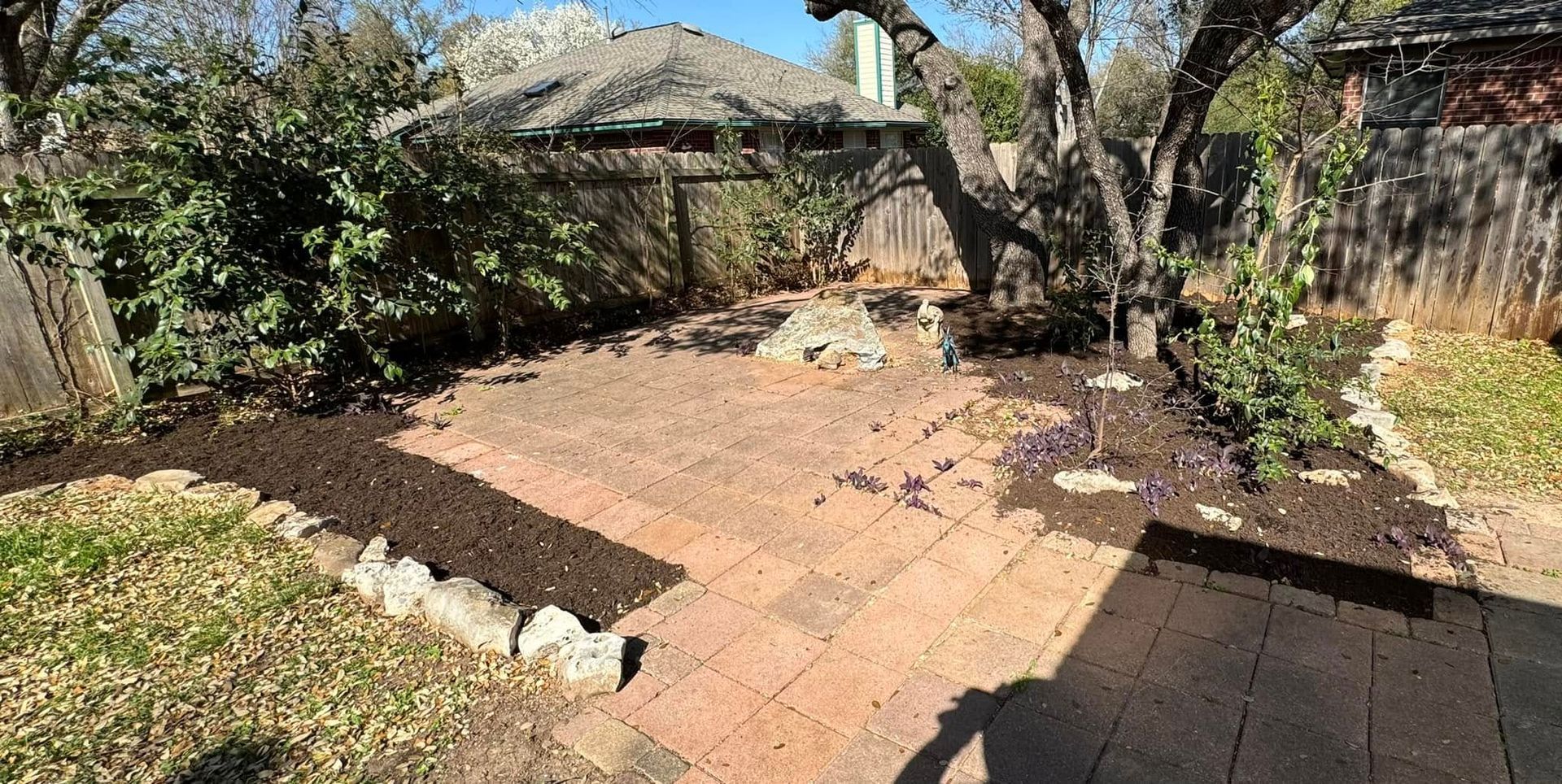 Backyard patio with brick pavers, mulch beds, tree, and wooden fence.