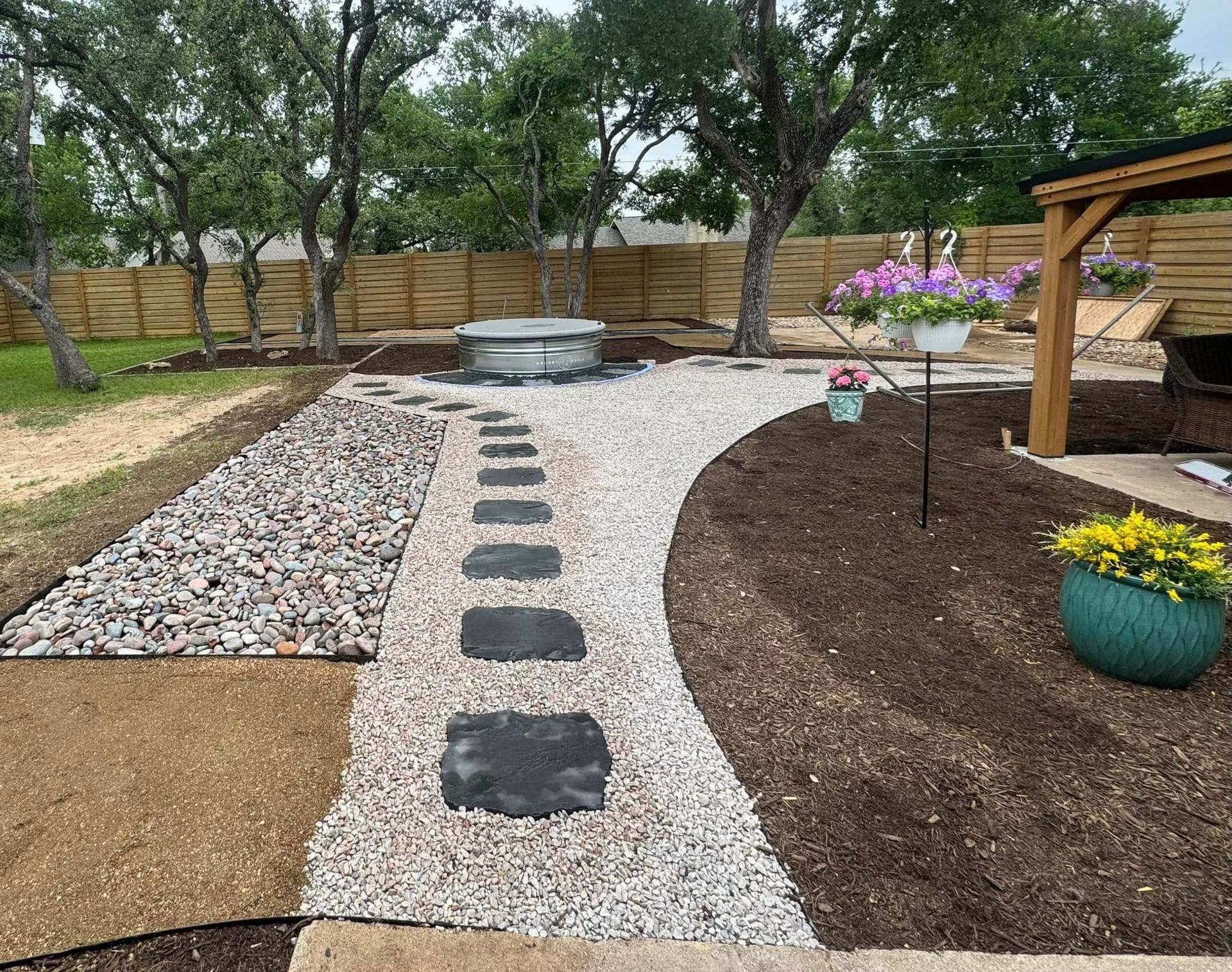 A backyard landscape with a stepping stone path, gravel beds, mulch, and a metal tub.