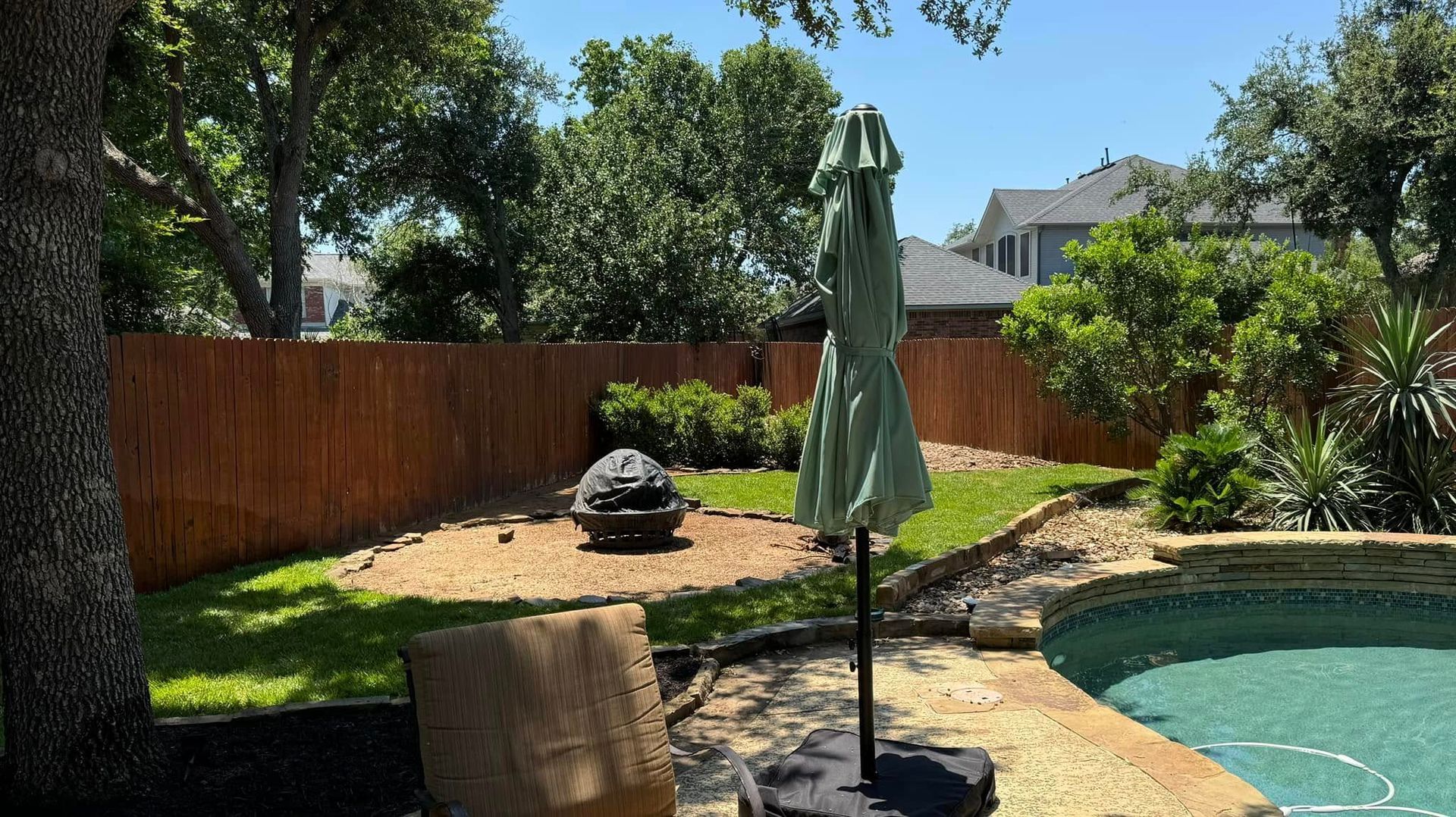 Backyard with a pool, lawn, and wooden fence on a sunny day. An umbrella stands near the pool.