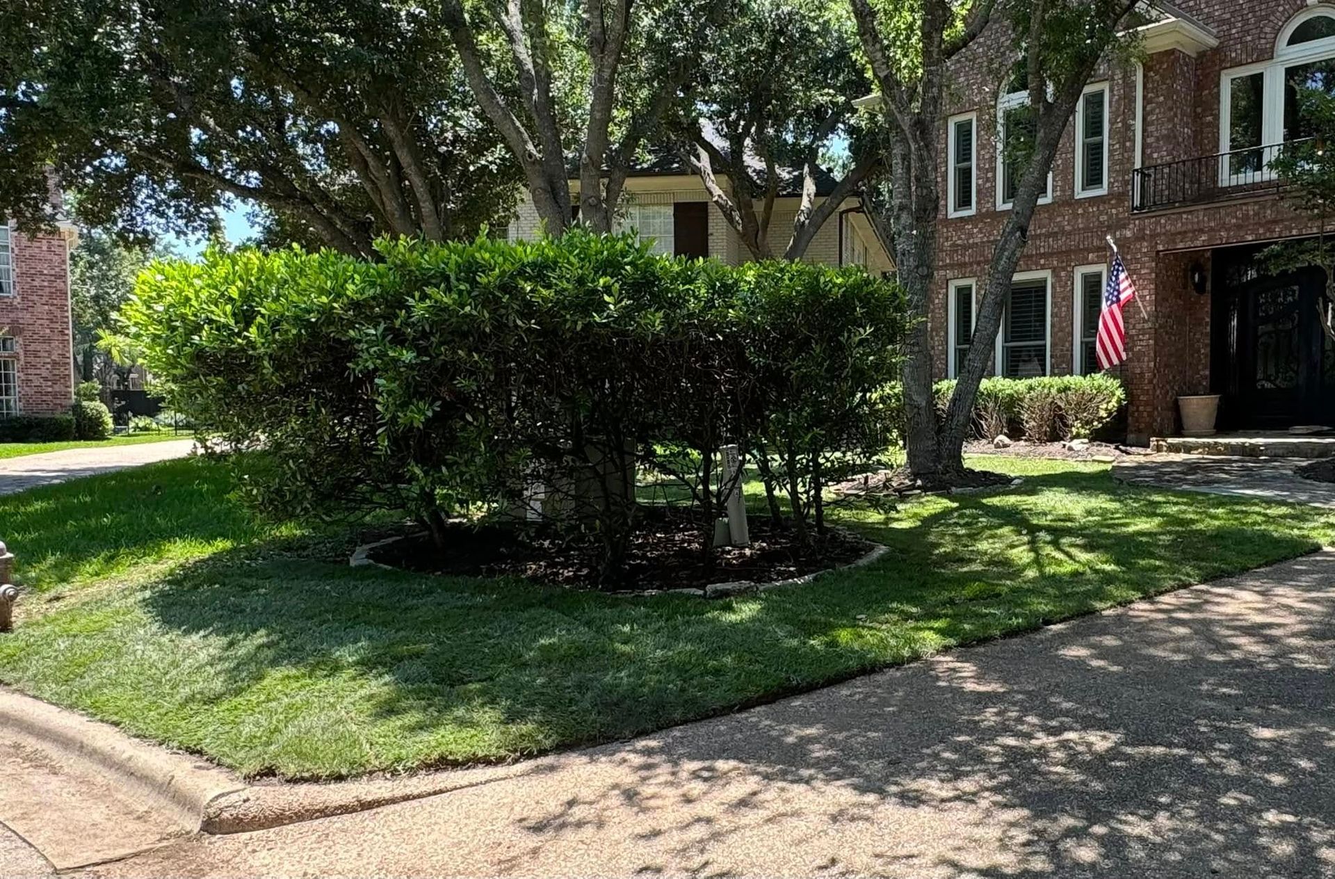 Lush green bushes and manicured lawn in front of a brick house with an American flag.