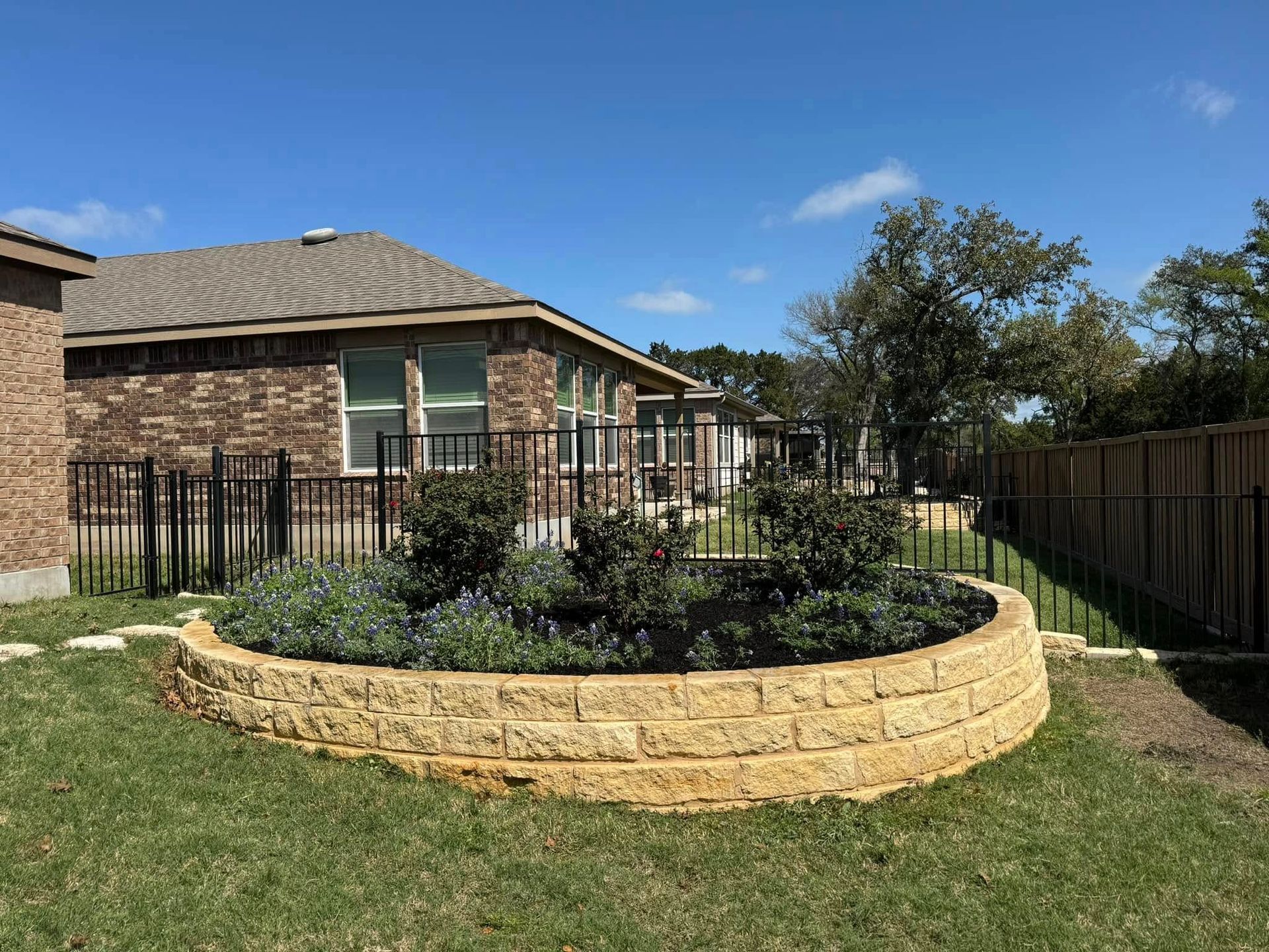Curved, raised garden bed with plants, brick house in background, black fence, and sunny blue sky.