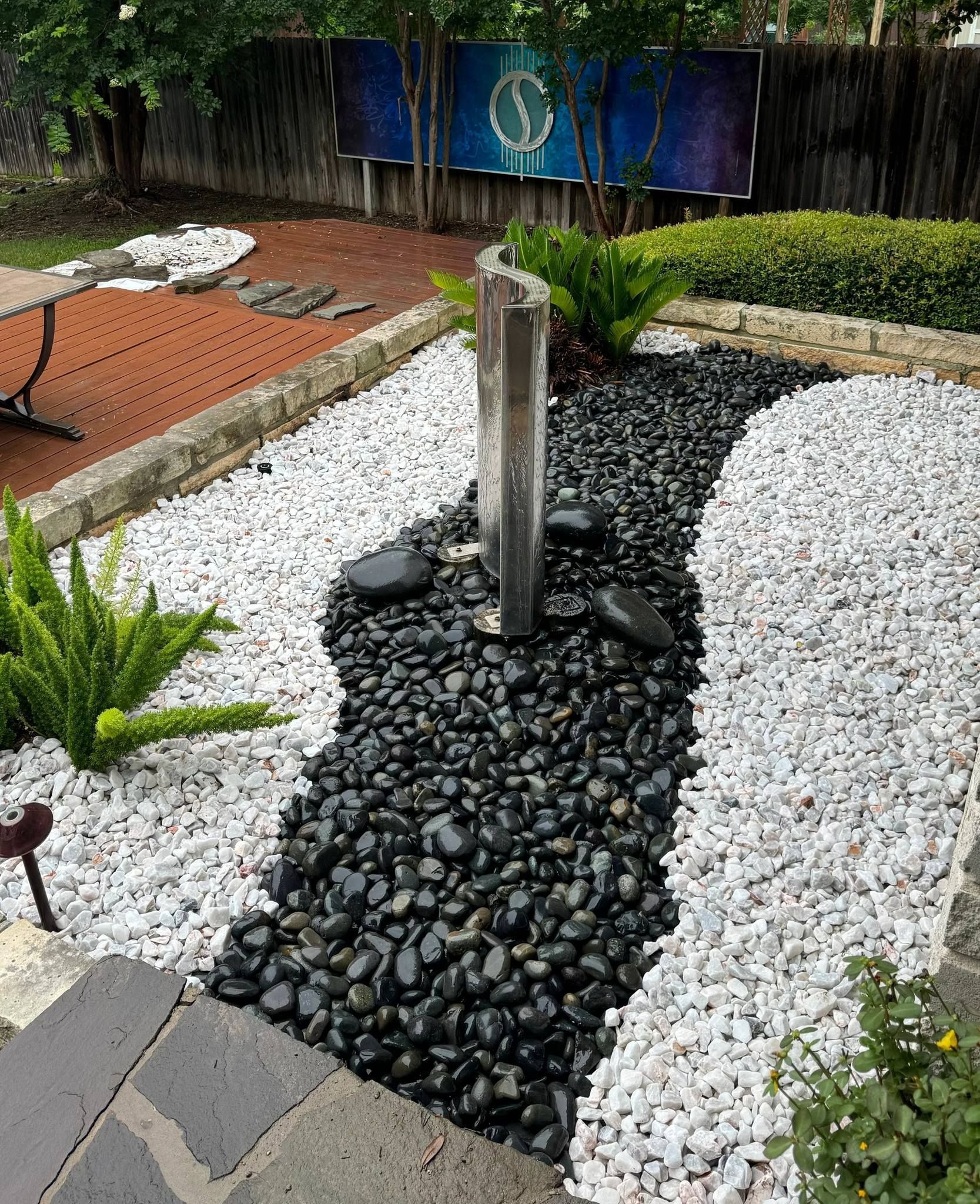 A modern garden with a stone fountain, black and white river rock pathways, and greenery.