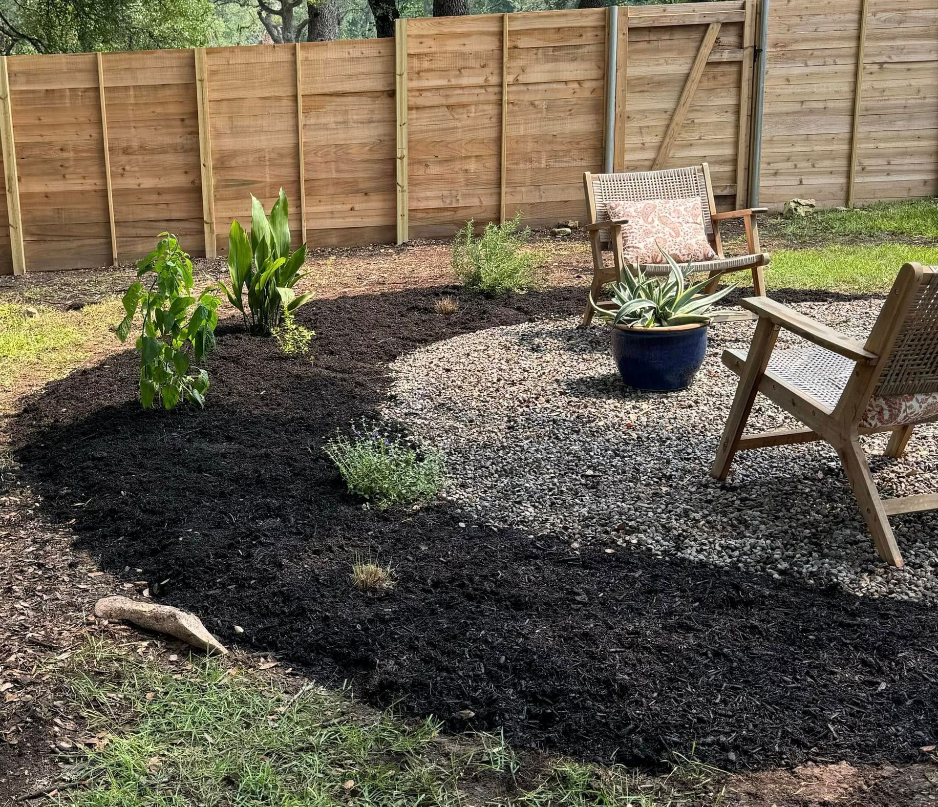 A backyard patio with a wooden fence, seating, plants in a mulch bed, and gravel.