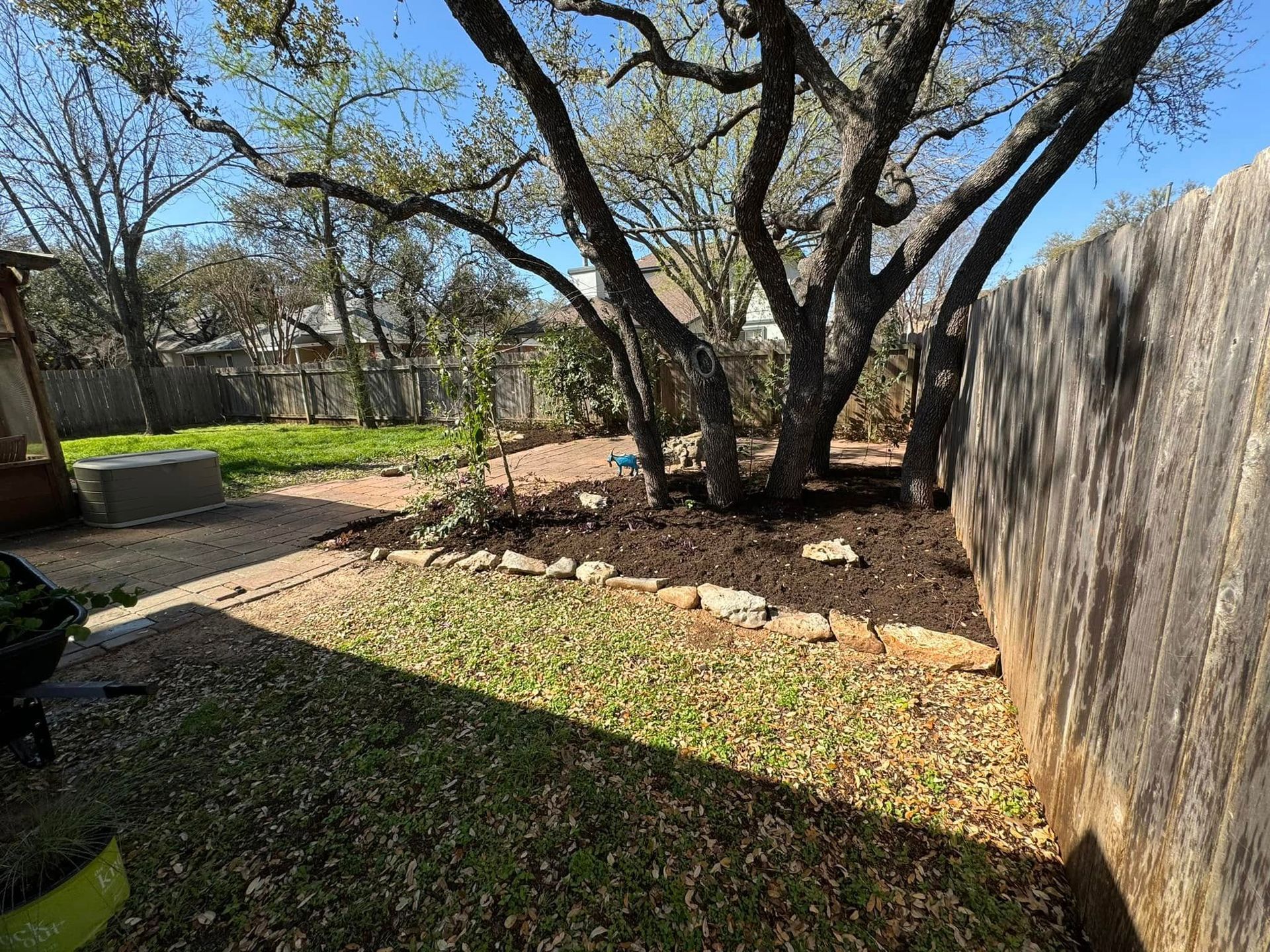 Backyard with a large tree surrounded by mulch, bordered by stone. A wooden fence runs along the right.