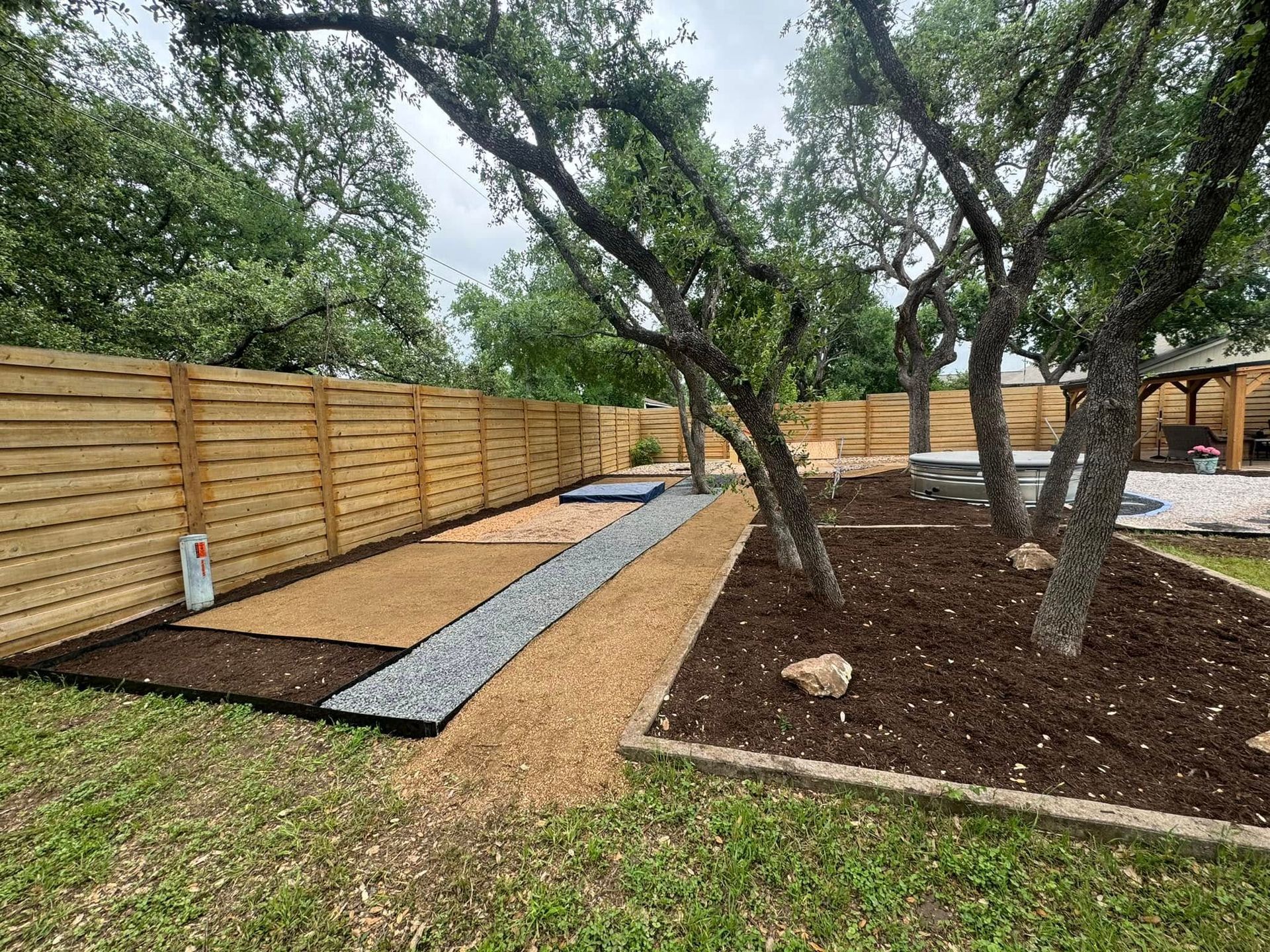 A backyard with a wooden fence, mulched flower beds, and gravel paths under large trees.