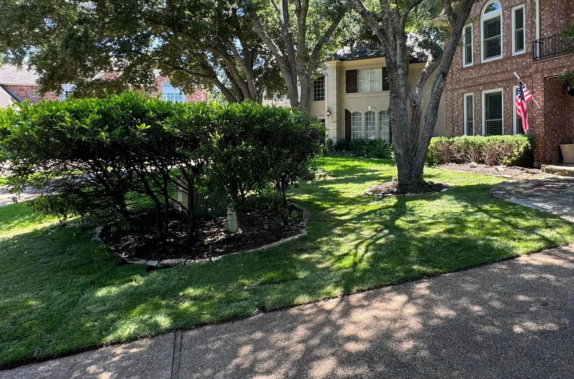 Green lawn and shrubs in front of two-story brick and beige houses; shade from a large tree.